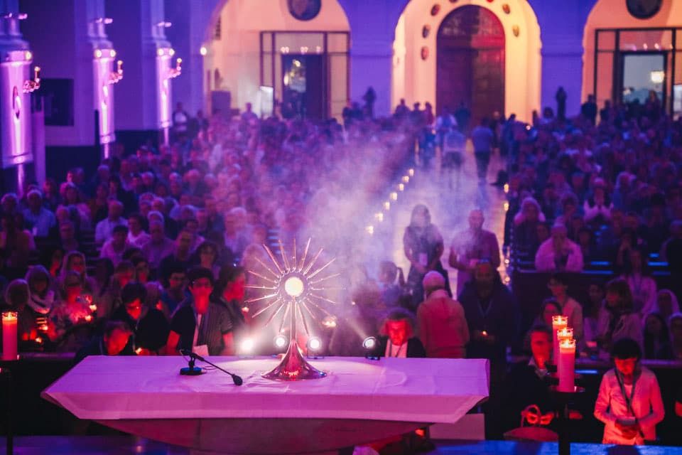 An illuminated monstrance stands on the altar. The church is full of people adorating the Blessed Sacrament. The light is warm and pleasant. The church is atmospherically lit