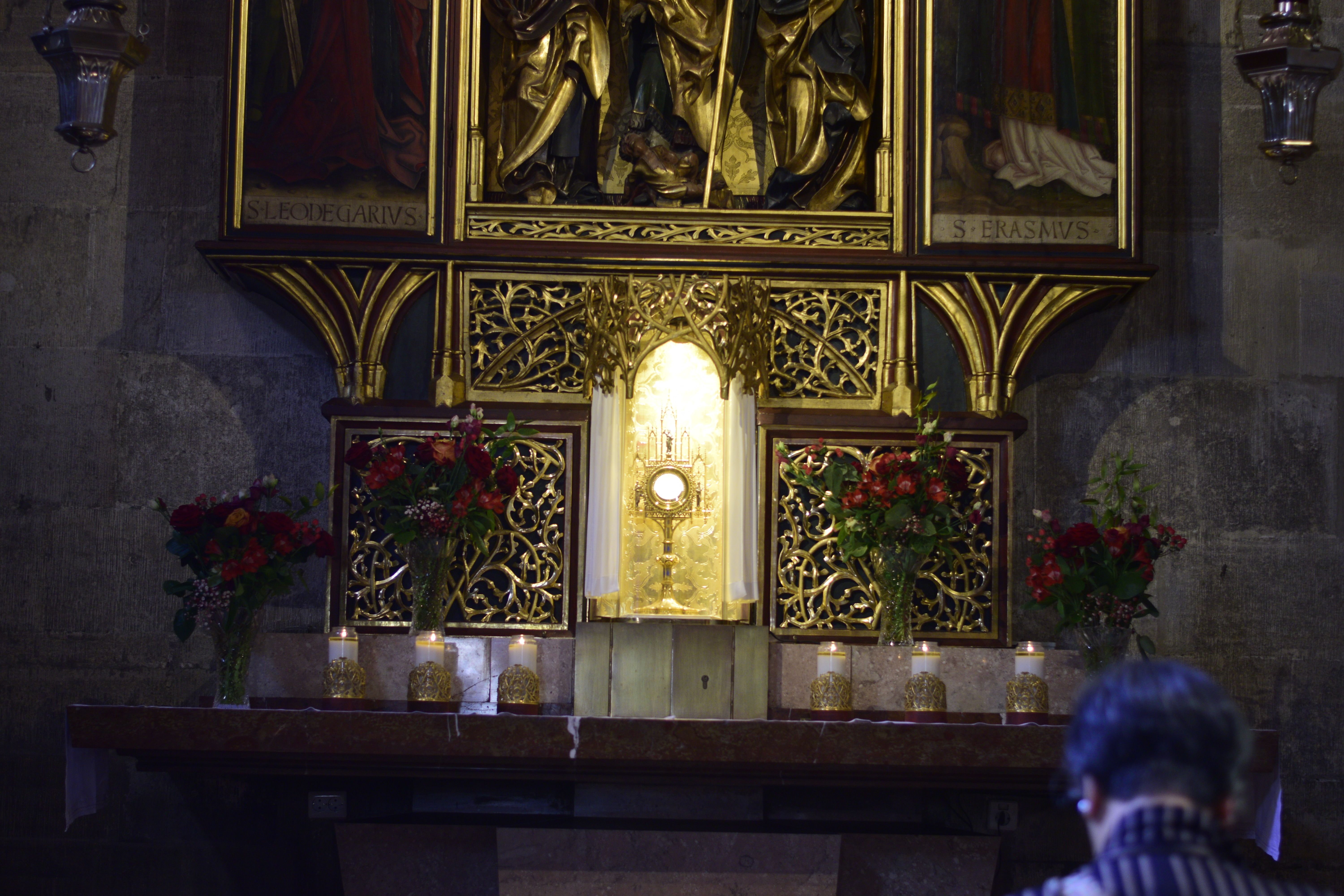 Gothic altar in a side chapel of St. Stephen's Cathedral, monstrance with the Blessed Sacrament shining; woman kneels in pew and prays