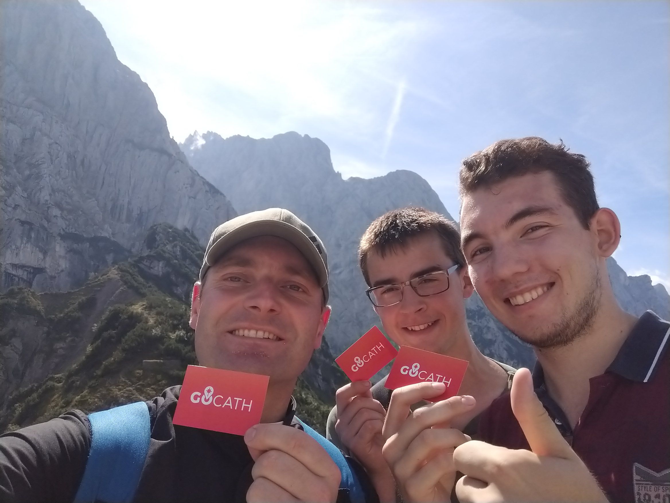 Priest and two young men are happy, are in the mountains, the sun is shining, all are holding an orange GOCATH business card in their hand