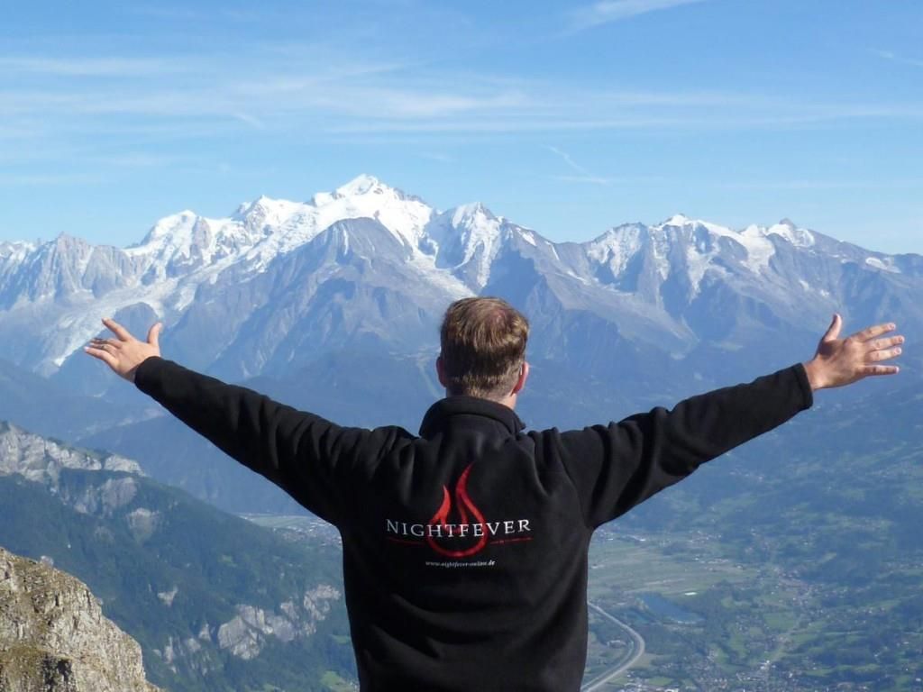 Man with open arms and Nightfever jacket, high snowy mountains in the background