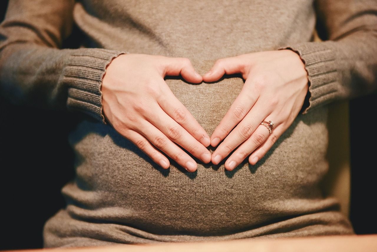 Pregnant woman in a brown wool dress touching her pregnant baby bump with her hands and forming a heart